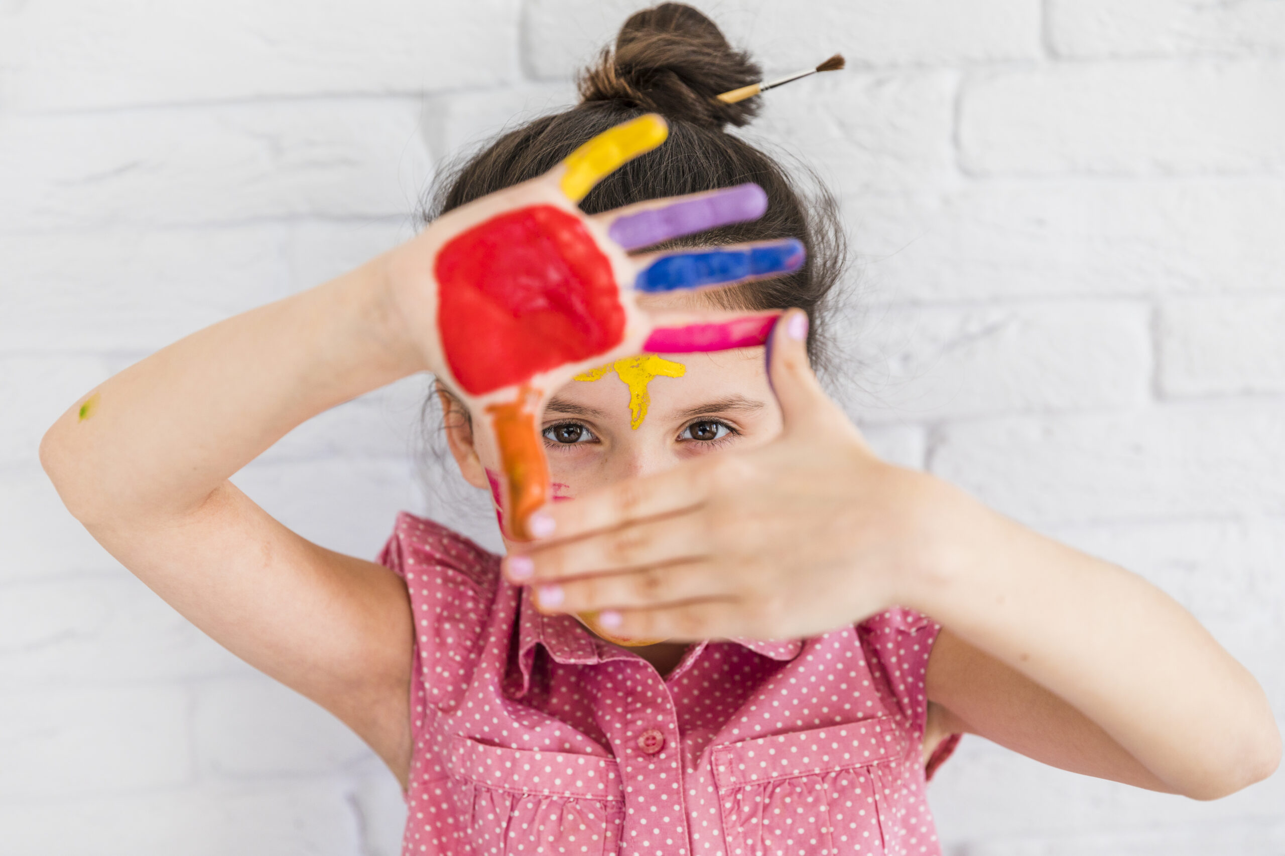 portrait girl looking through her painted hands standing against white brick wall scaled